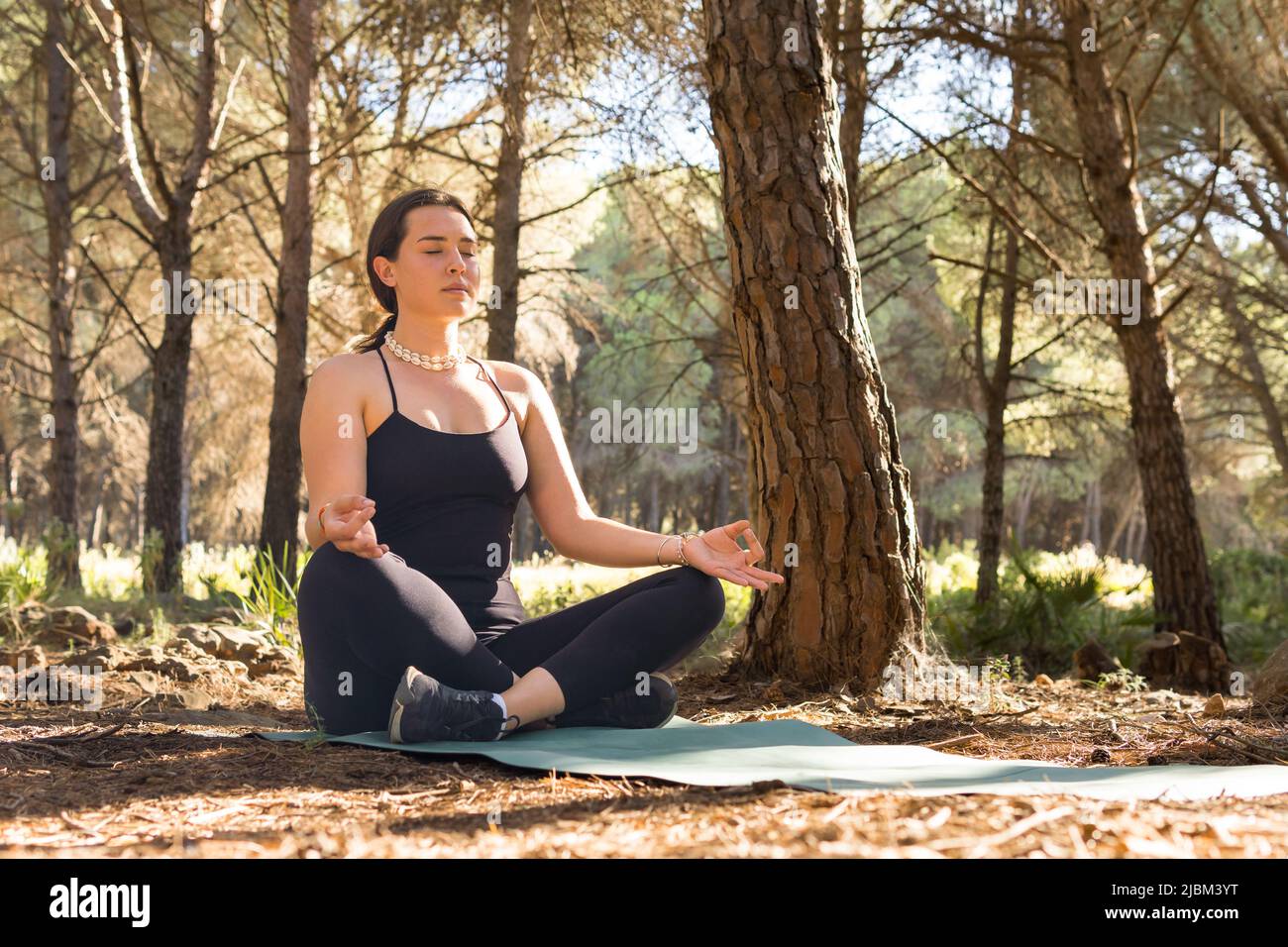 beautiful young adult girl doing meditation in the lotus posture at sunset in a forest surrounded by trees. open plane, horizontal. Stock Photo