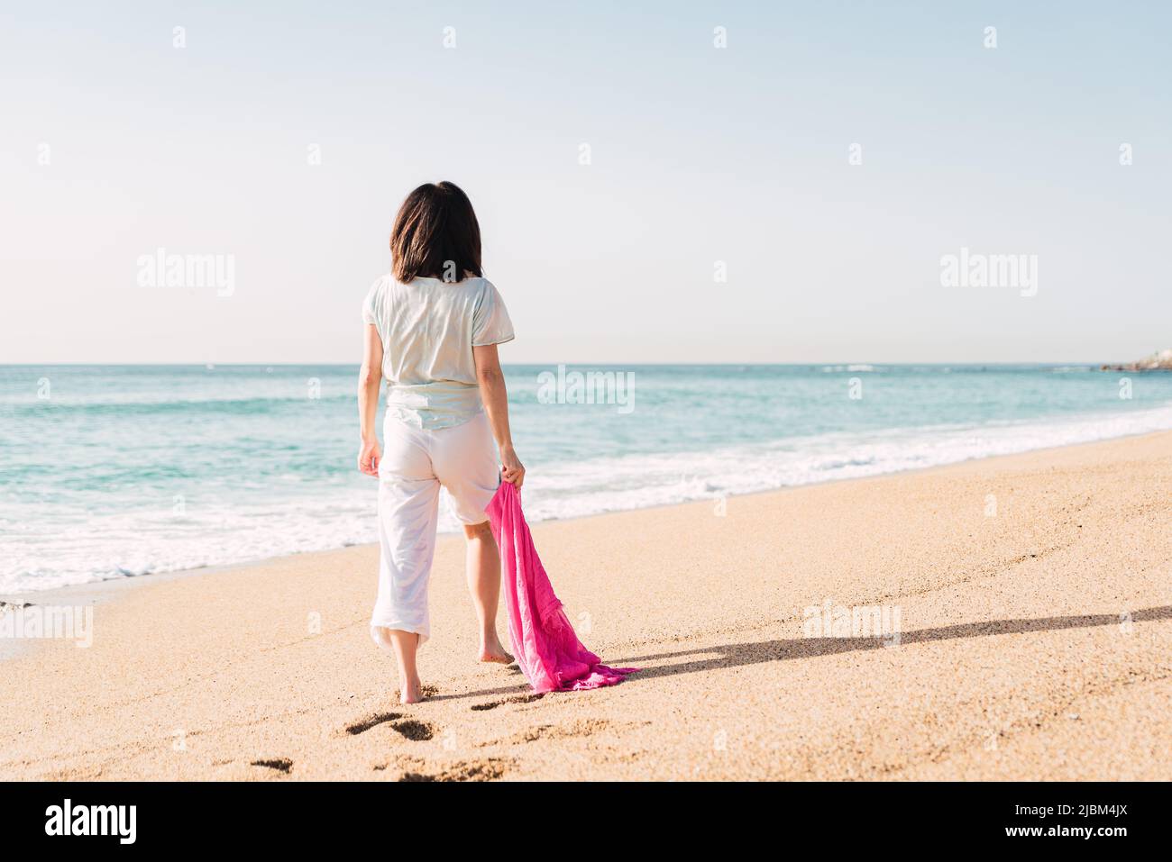 Back view of unrecognizable female in white clothes and with scarf having stroll on sandy coast near sea in sunny weather Stock Photo
