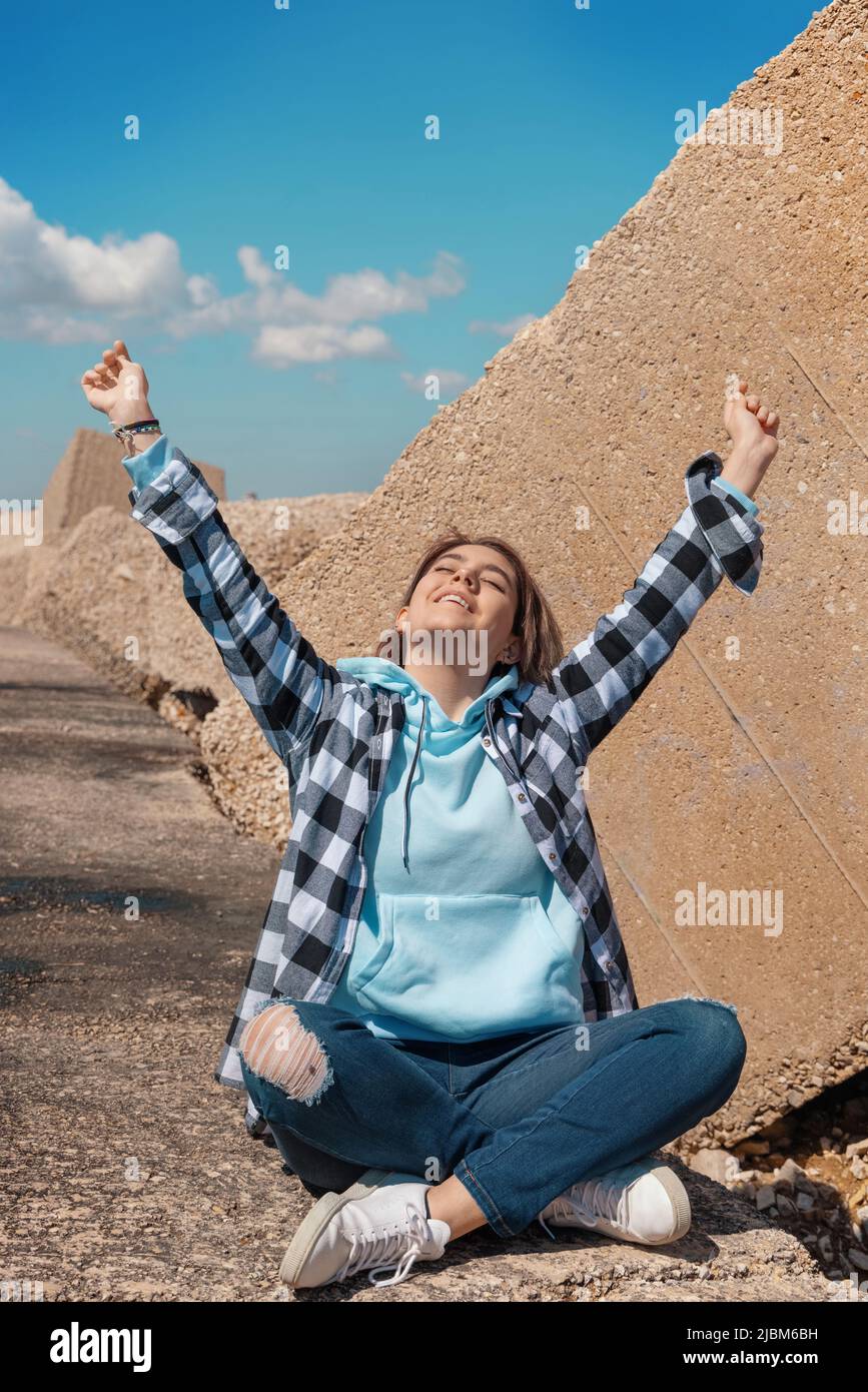 Happy Teenager girl in light blue hoodie and plaid shirt raising arms overhead. Smiling Teen girl sitting outdoor cross-legged and enjoying the sunny Stock Photo