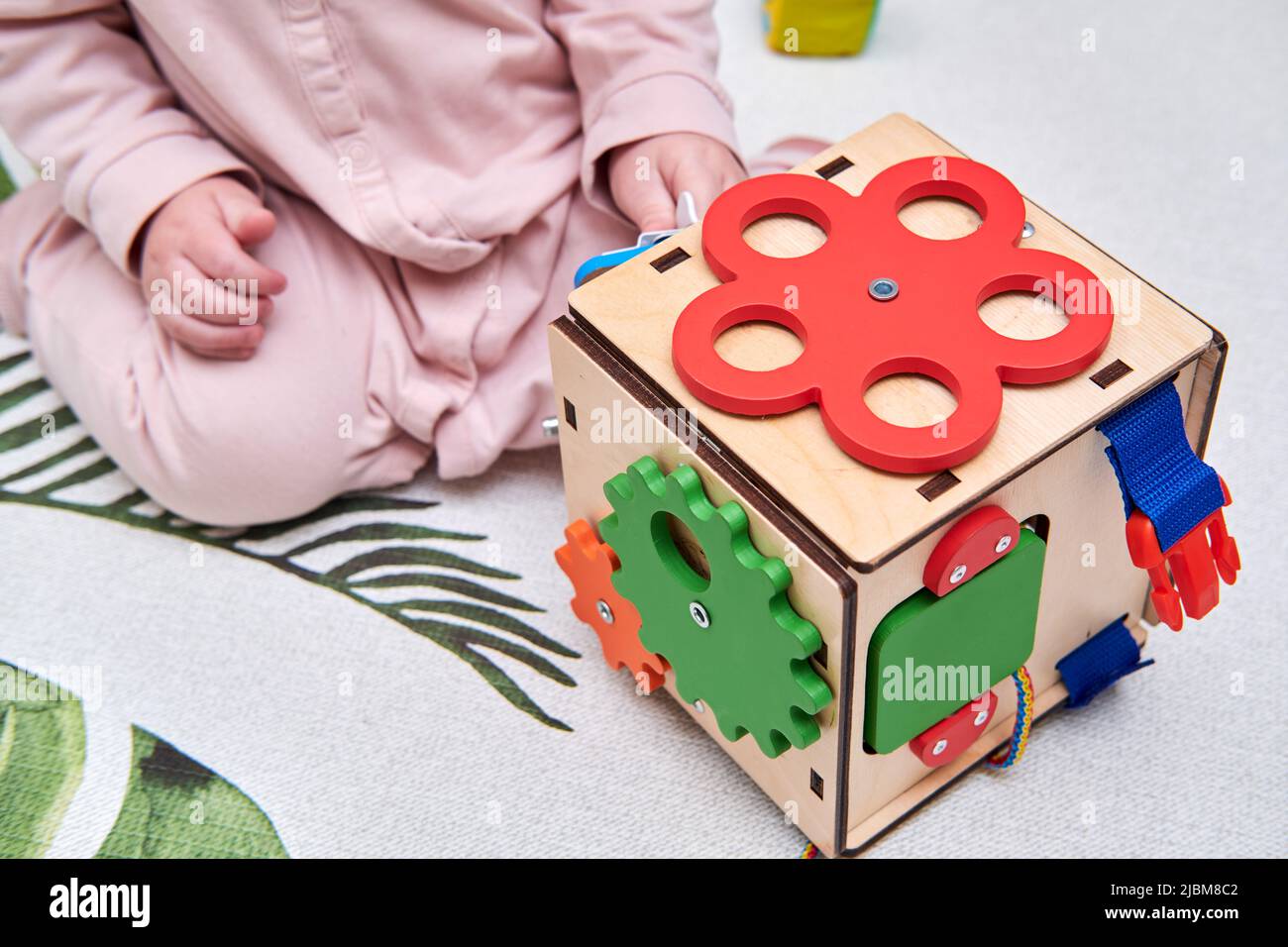 Happy toddler baby boy is playing with a busyboard cube on the nursery floor. Educational toys for children, wooden game panel. A child aged one year Stock Photo