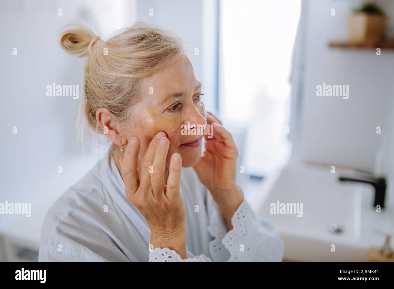 Beautiful senior woman in bathrobe, applying eye patches for puffiness while looking in the mirror Stock Photo