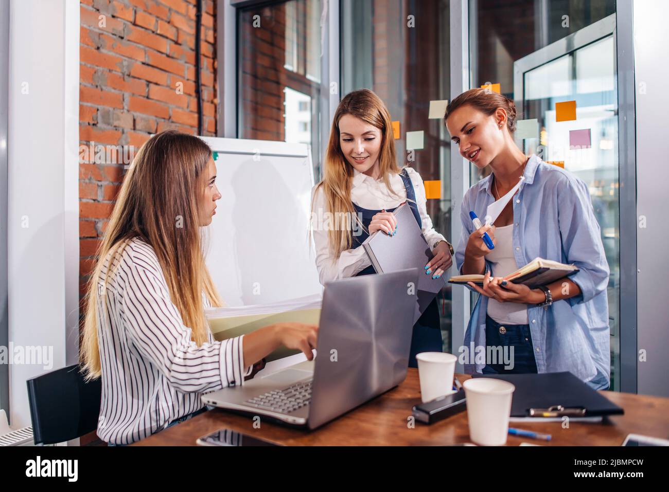 Young female manager sitting at desk pointing at laptop explaining giving tasks to her employees standing writing the information in notebooks in offi Stock Photo