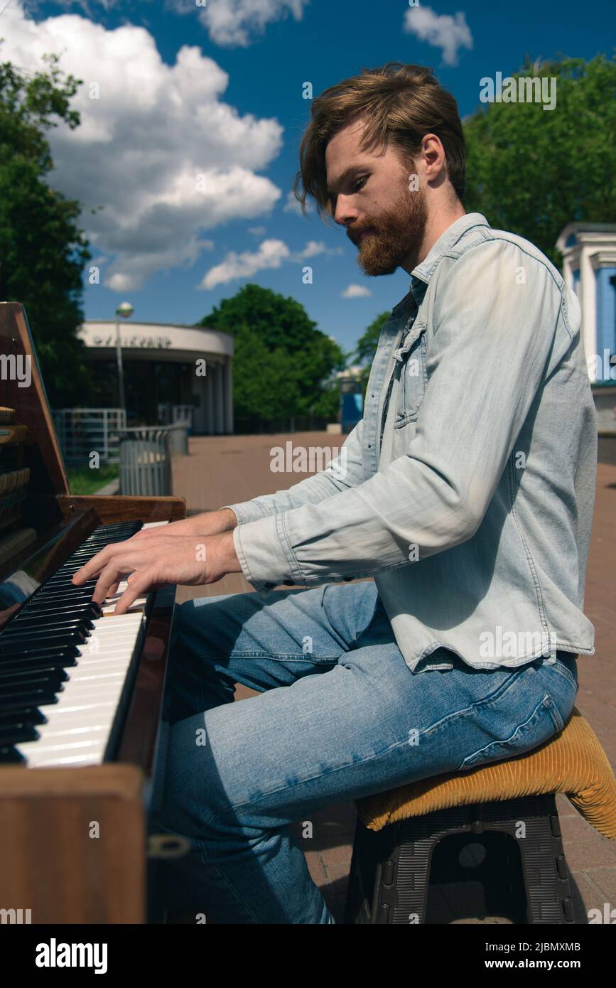 Street musician playing old piano in downtown. Young bearded man pianist playing music outdoors. Handsome talented busker Stock Photo