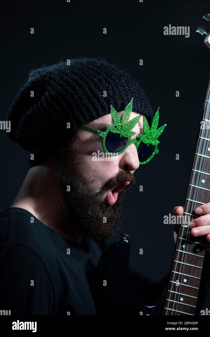 portrait of a bearded musician guy, wearing marijuana sunglasses, emotionally playing the electric guitar. on a dark background Stock Photo