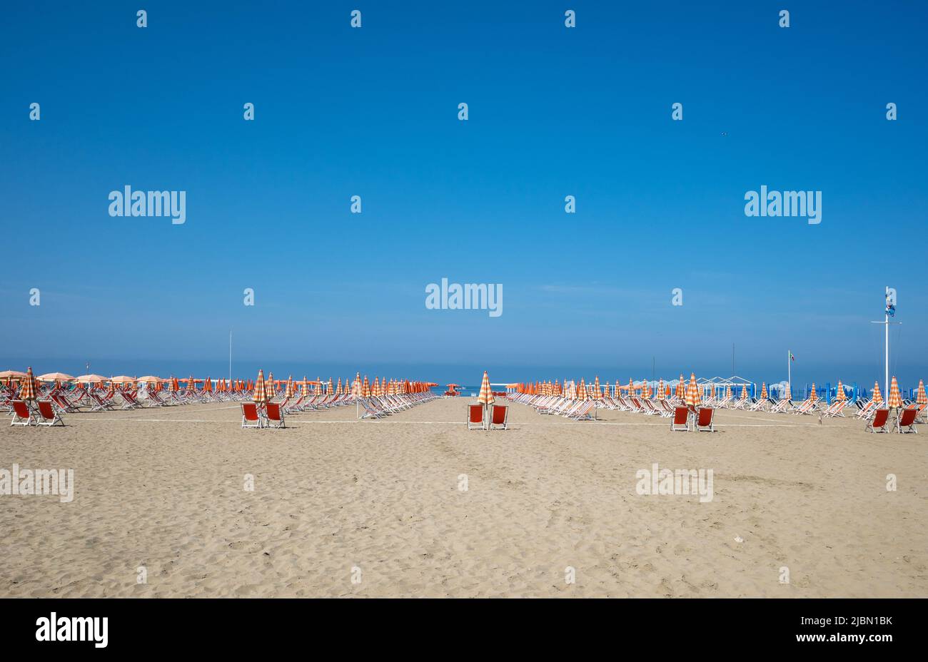 A view of red beach deck chairswith orange and white umbrellas on a sunny day with a clear blue sky. Stock Photo