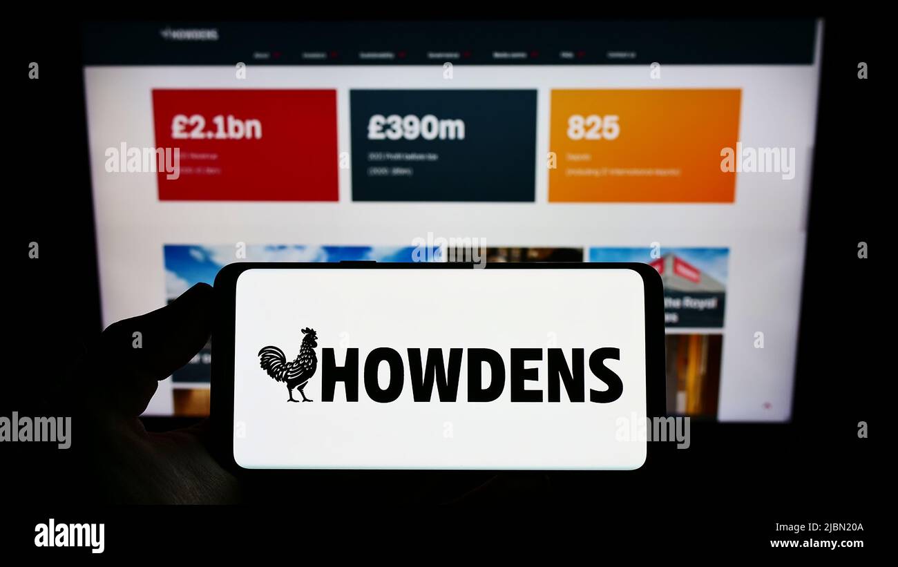 Person holding smartphone with logo of British company Howden Joinery Group plc on screen in front of website. Focus on phone display. Stock Photo