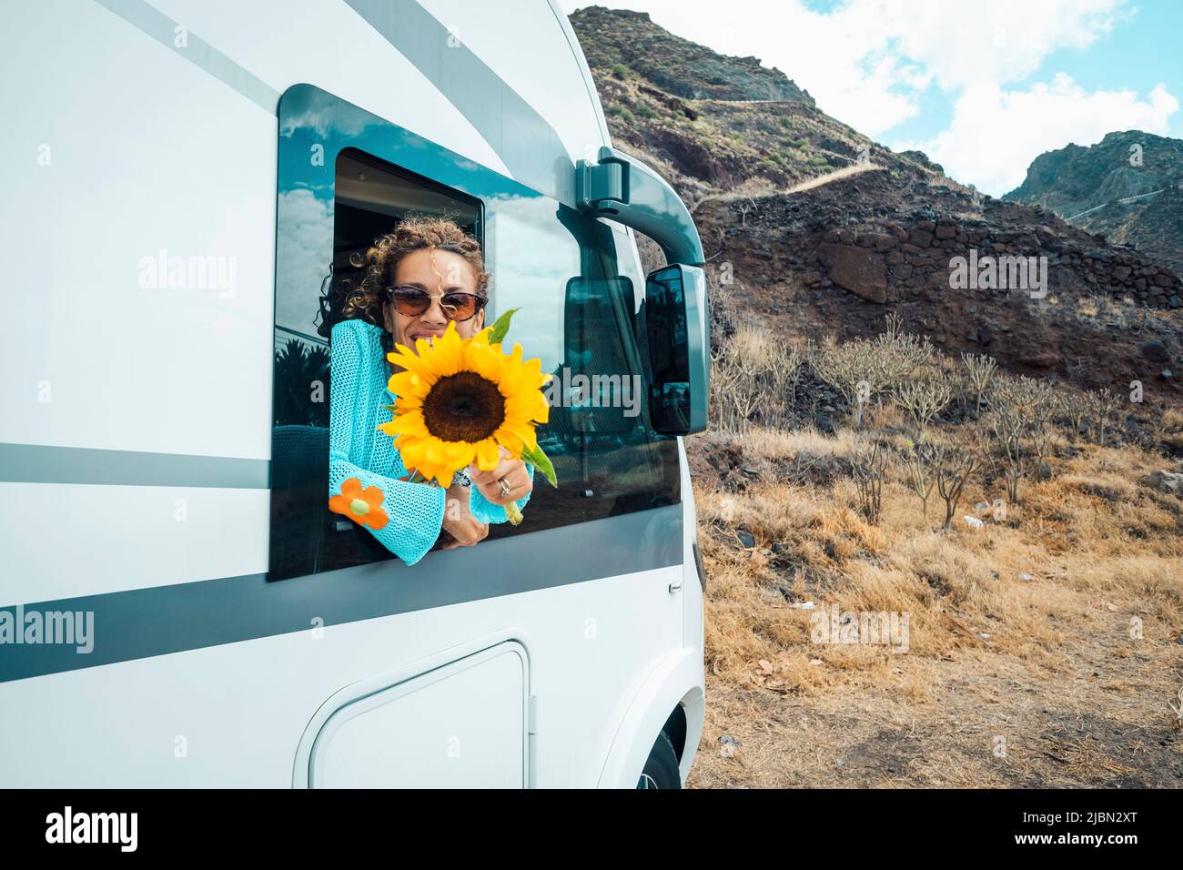 Modern hippy travel lifestyle. Happy young woman out of the camper van window enjoy trip destination and holding big sunflower. Concept of rent rv veh Stock Photo