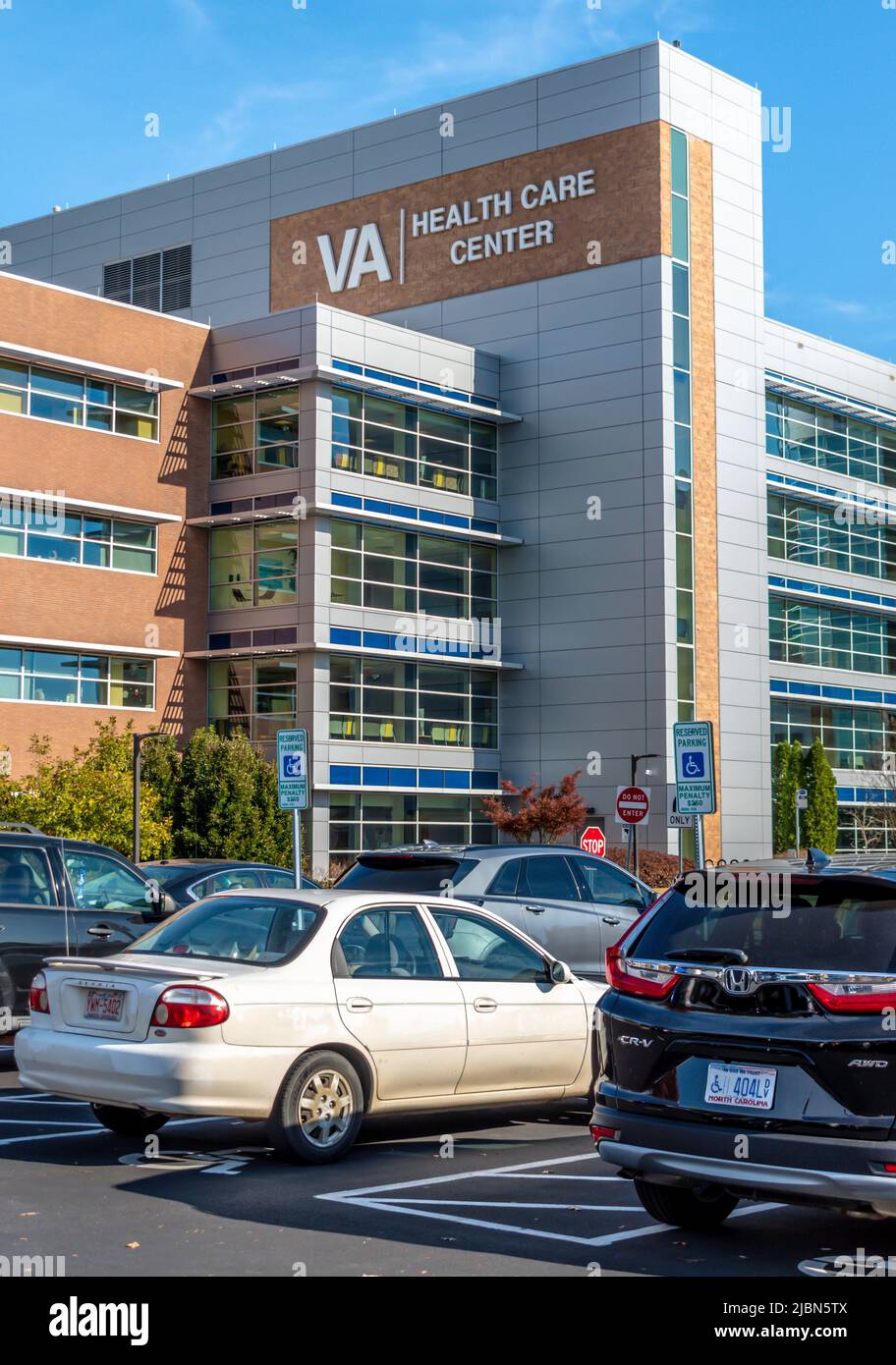 VA Health Care Center's exterior facade brand and logo signage on a bright sunny day with blue sky, hazy clouds, floors, windows, and parked vehicles. Stock Photo
