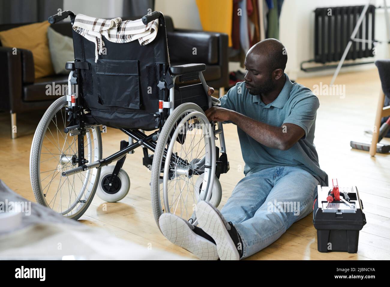 African man with disability sitting on floor and fixing his broken wheelchair with instruments Stock Photo