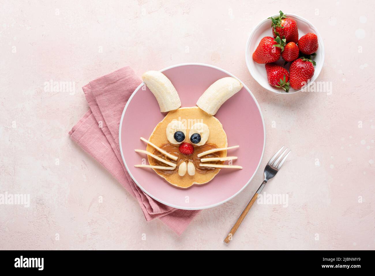 Funny cute bunny pancake art for kids for breakfast. Pancake with fruits shaped as rabbit or bunny on pink background, top view Stock Photo