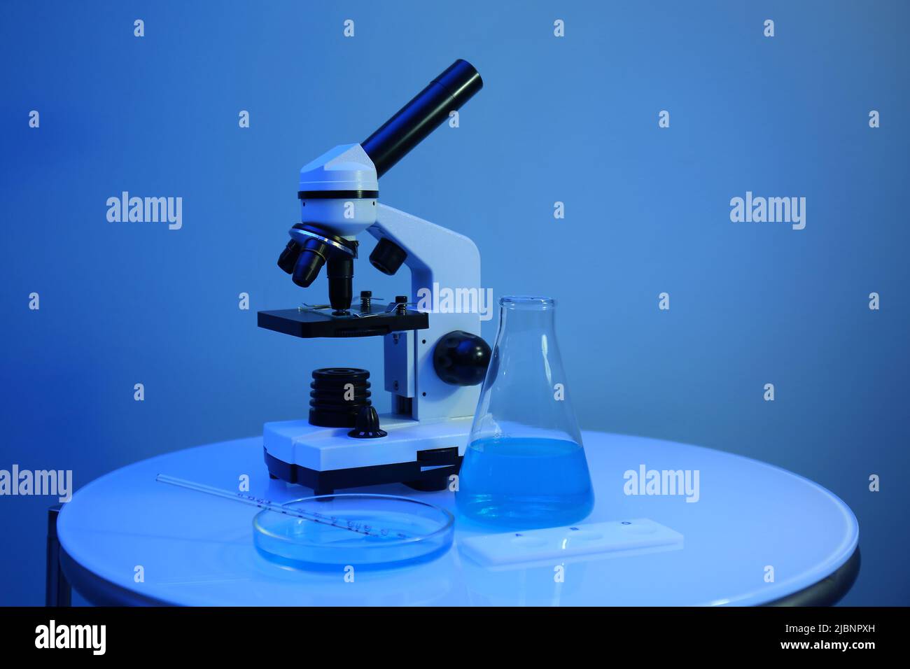 Different laboratory glassware with samples and microscope on table near blue wall Stock Photo