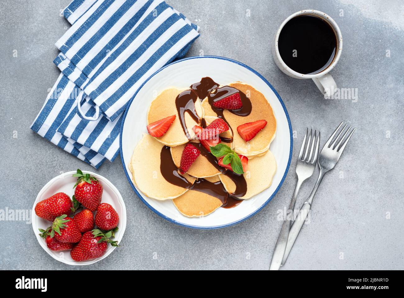 Crepes or pancakes with strawberries and chocolate on plate served with cup of black coffee, grey concrete table background. Top view Stock Photo
