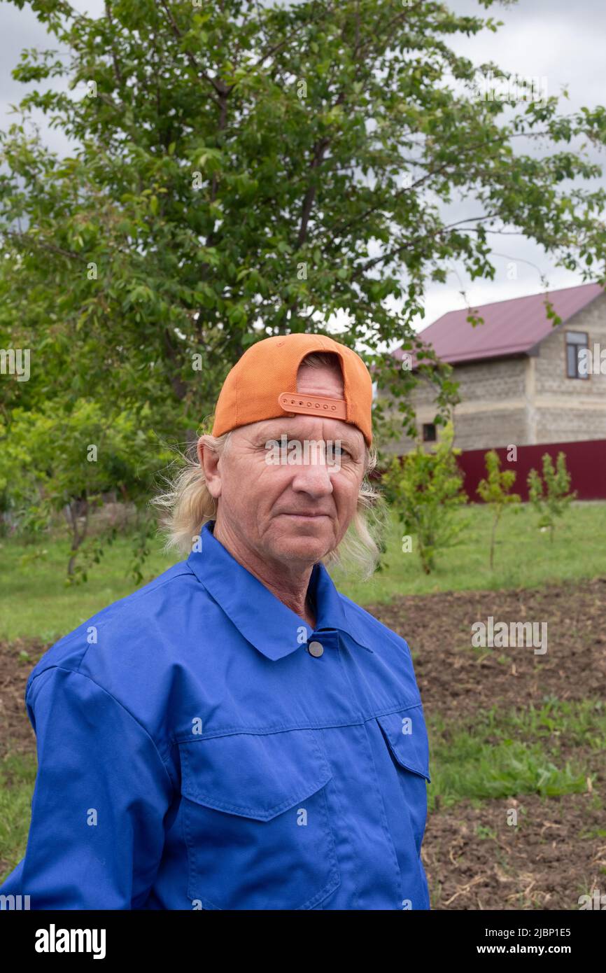 Mature male farmer in a cap on the background of the house and garden. Farming and growing plants. Stock Photo