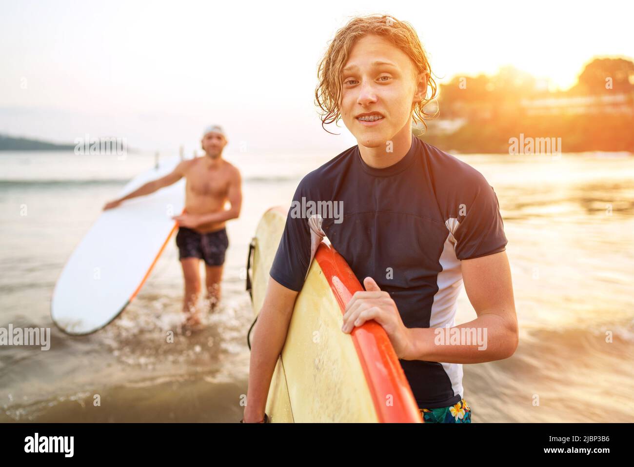Young teenager blond boy with a father carrying surfboards as they walking ocean sandy beach after surfing with beautiful sunset background. They are Stock Photo
