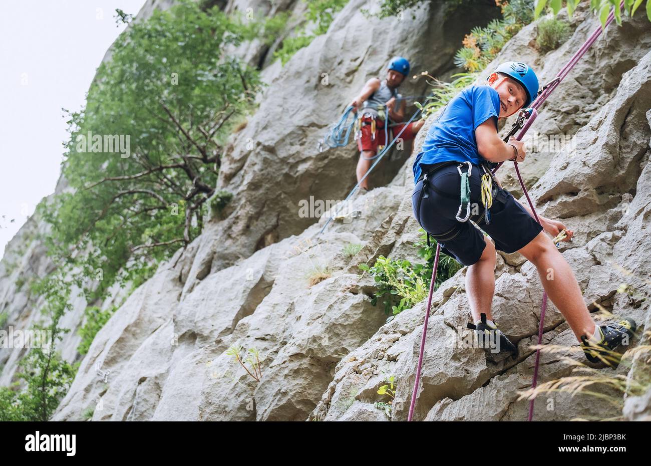 Smiling teen boy in protective helmet abseiling from cliff rock wall using rope, Belay device and climbing harness with father in Paklenica Park Croat Stock Photo