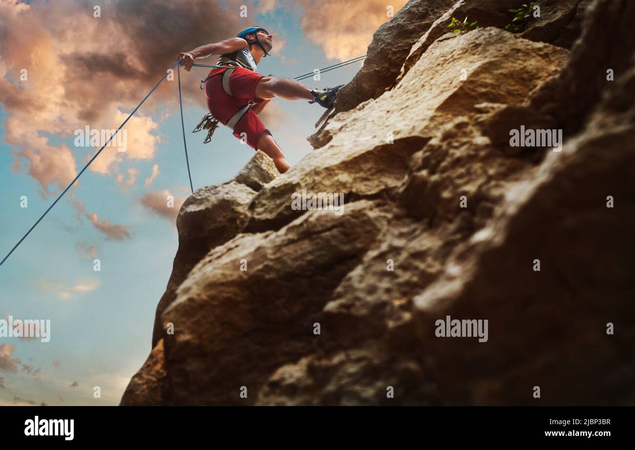 Muscular climber man in protective helmet abseiling from cliff rock wall using rope Belay device and climbing harness on evening sunset sky background Stock Photo