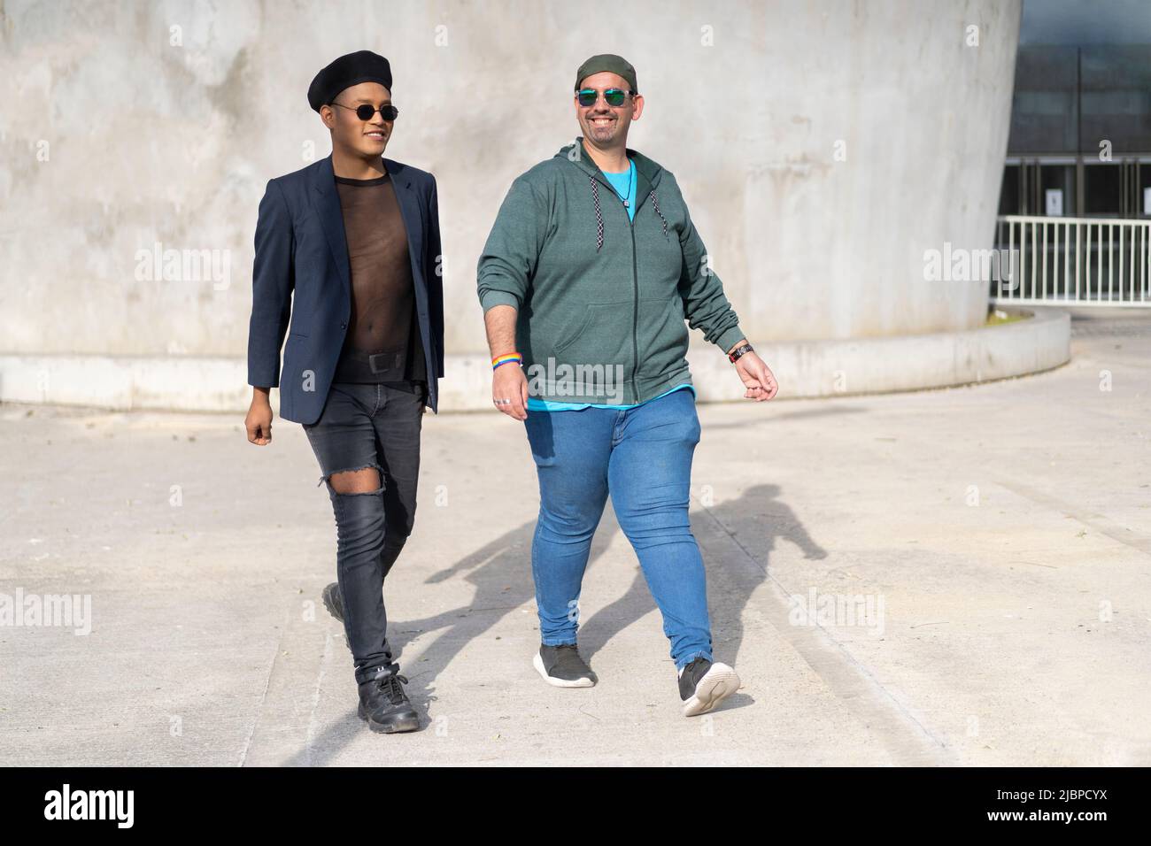 Gay Latino male couple walking in a park, wearing fashionable hats and sunglasses, smiling Stock Photo