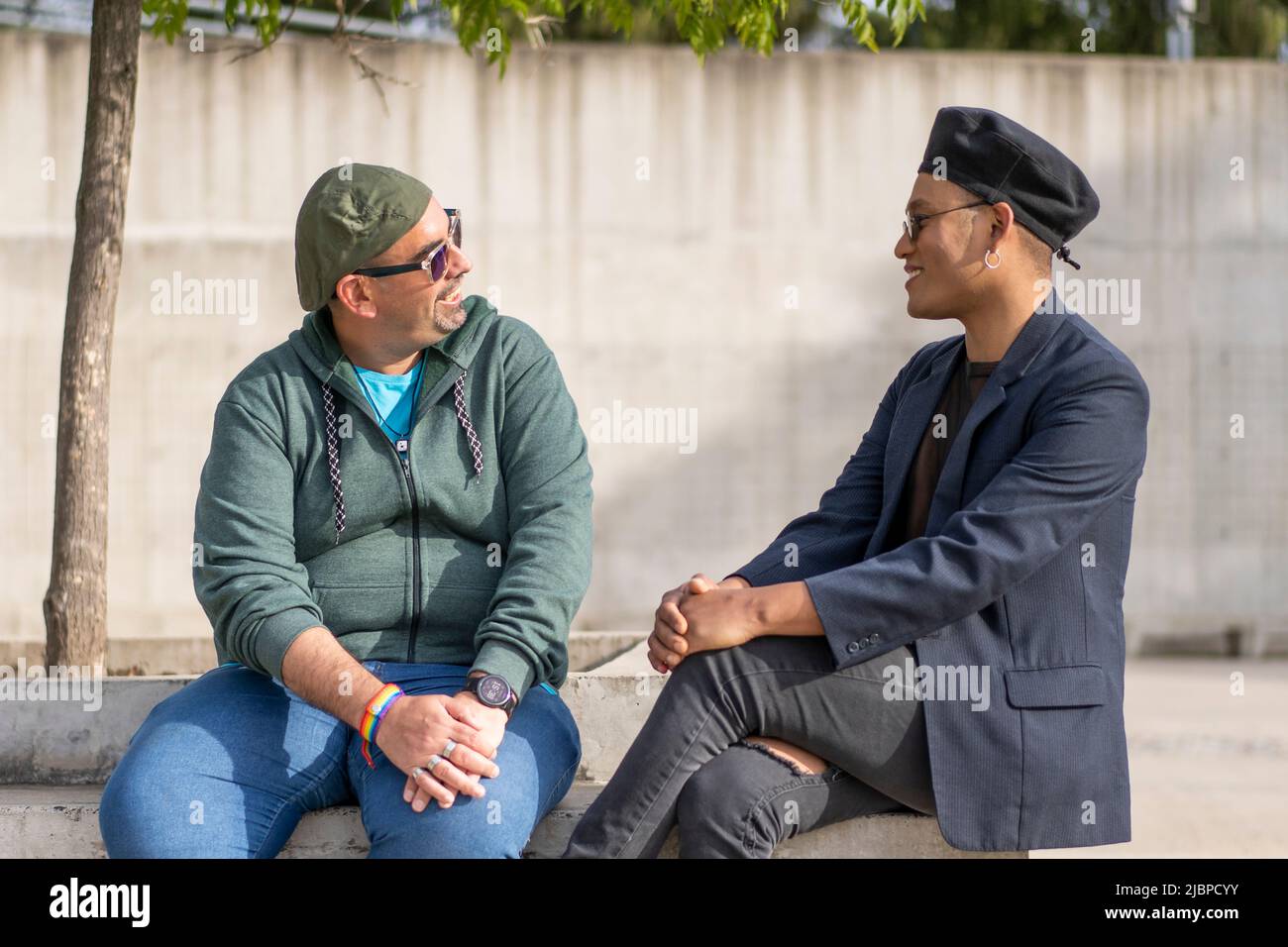 Gay Latino male couple sitting on a bench in a park, wearing fashionable hats and sunglasses, looking at each other smiling Stock Photo