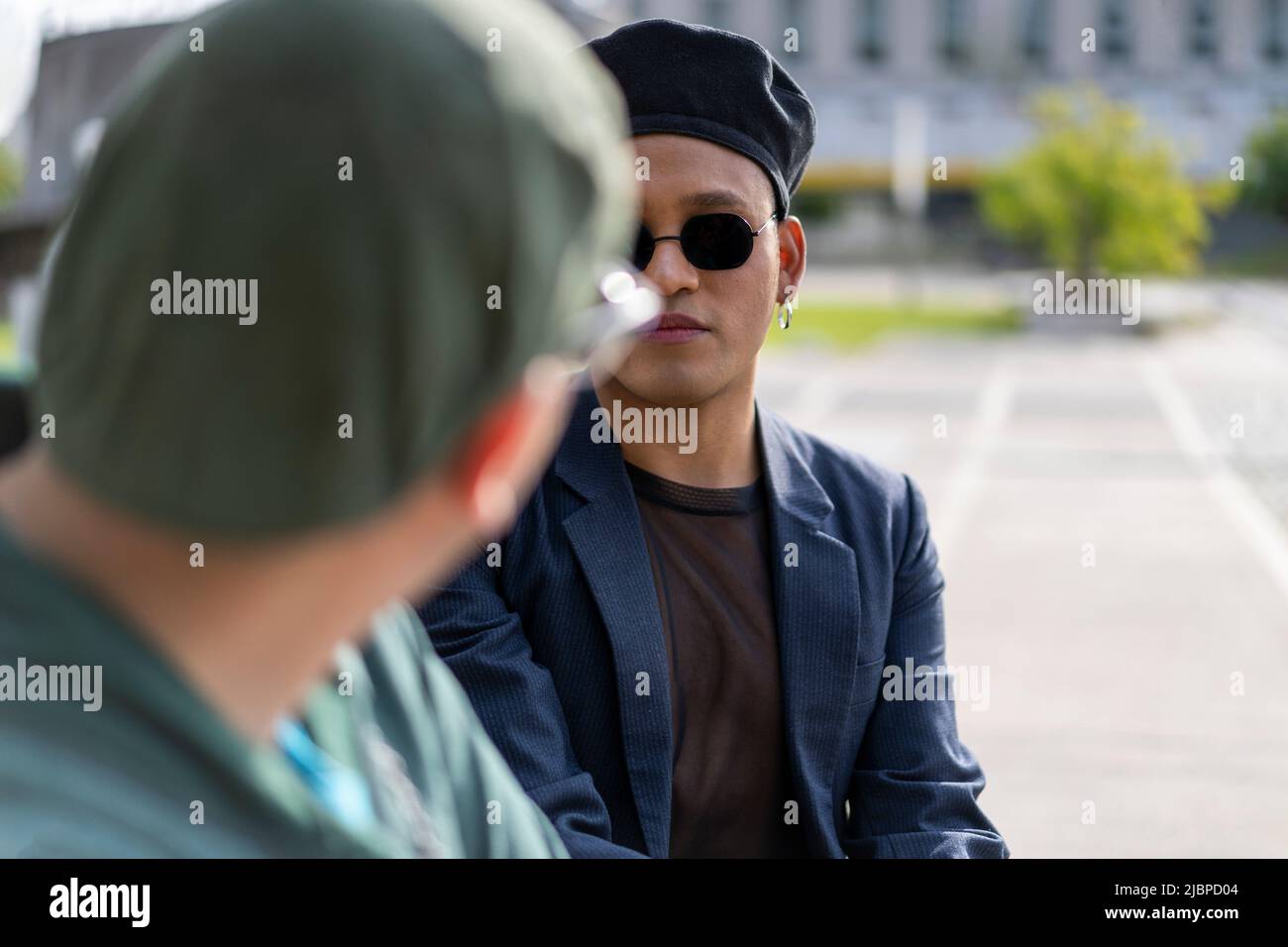Gay Latino male couple sitting on a bench in a park, wearing fashionable hats and sunglasses, looking at each other Stock Photo
