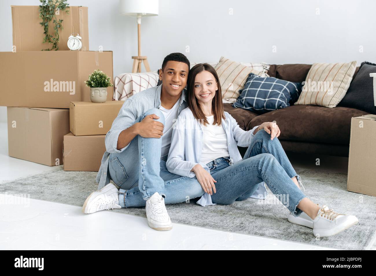 Happy multiracial family couple in love, hispanic guy and caucasian girl, sit on the floor in the living room of their new home, between cardboard boxes with household items, look at the camera, smile Stock Photo