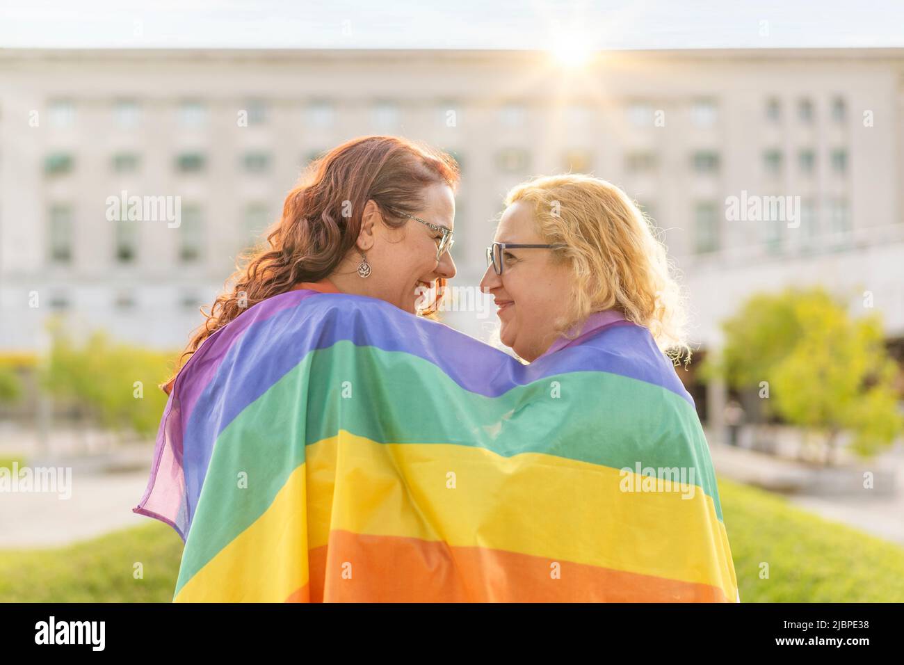 Lesbian women couple in love embracing each other, wrapped in a gay flag, in a park at sunset, with reflections of the sun. Concept of diversity, prid Stock Photo