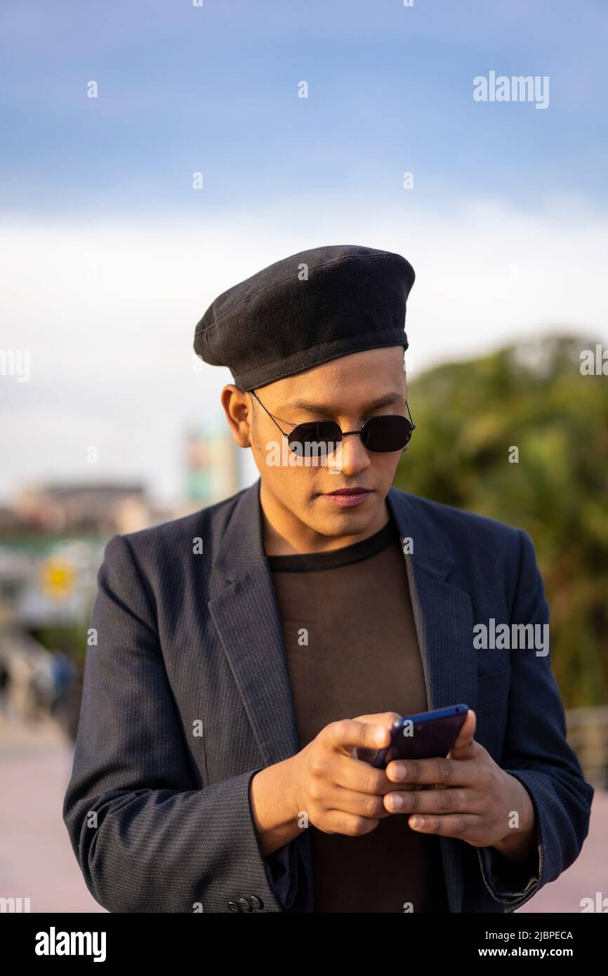 Latino gay male with makeup on wearing trendy hat looking at cell phone in a park Stock Photo