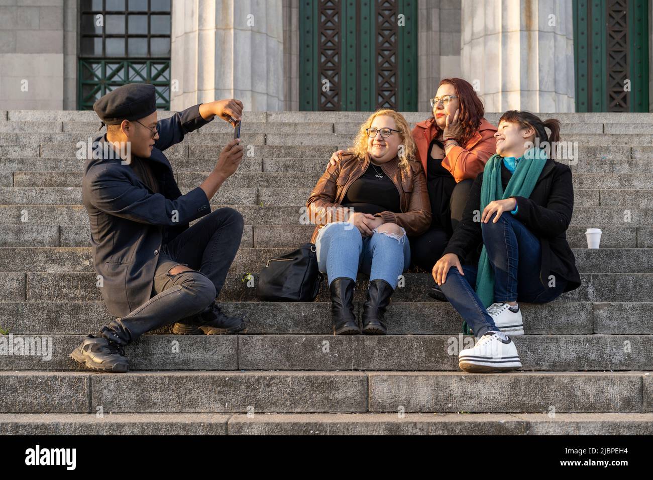 Multi ethnic group of friends sitting on some stairs having a good time chatting, laughing and taking photos Stock Photo