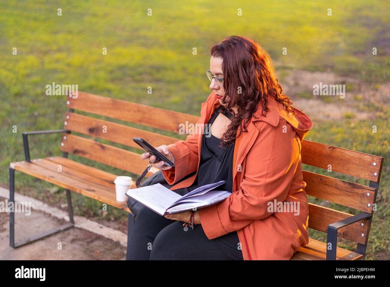 Woman sitting on a bench in a park checking her agenda and cell phone while having a coffee Stock Photo