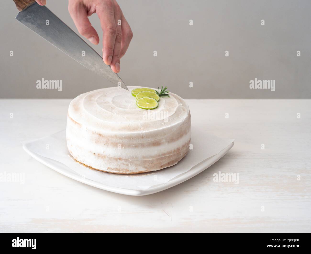 A Man's Hand Cuts a Vegan Birthday Cake with Almond, Coconut and Lemon Base with a Large Knife on a White Table and a White Background Stock Photo