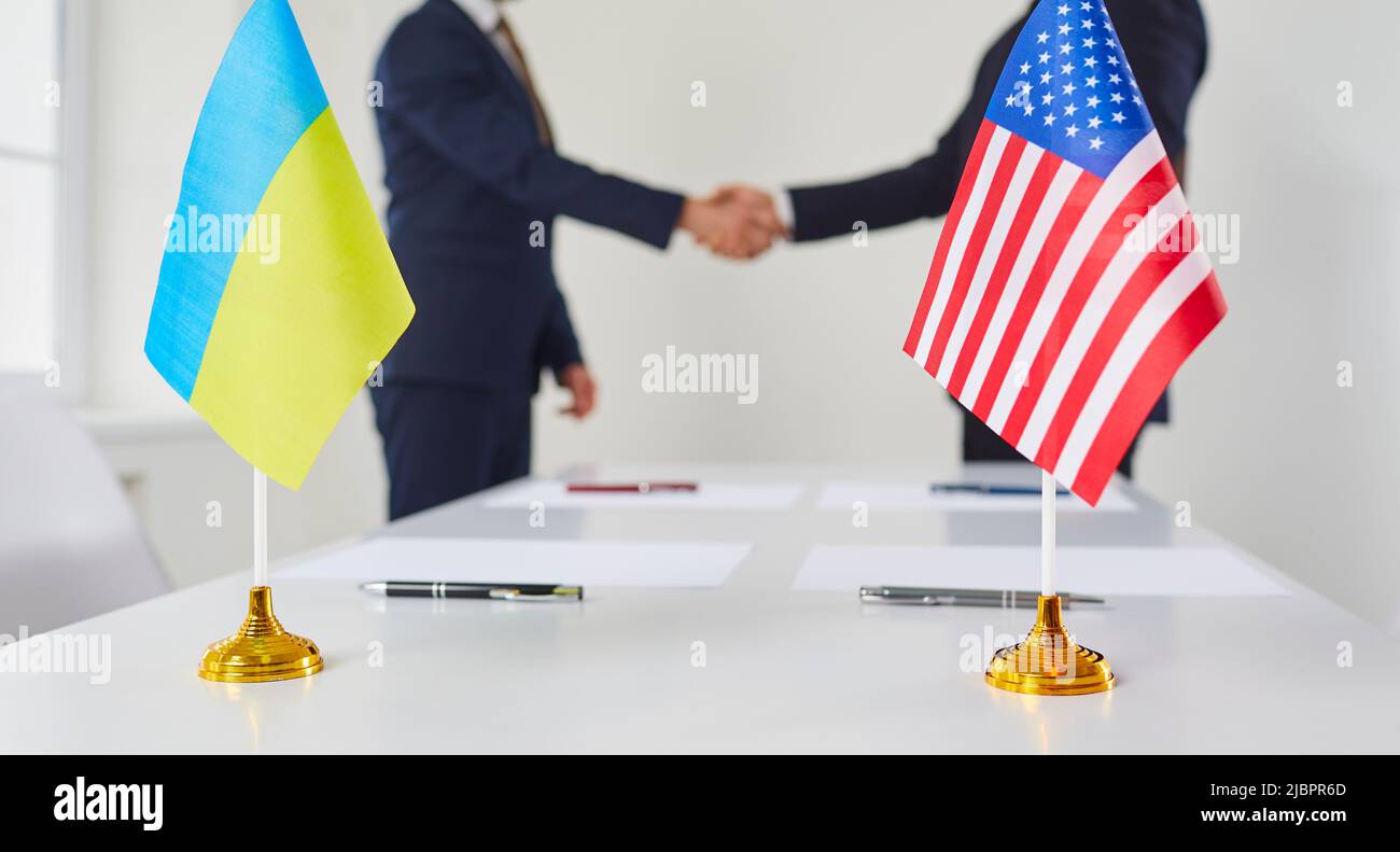 Small flags of Ukraine and America stand on table against background of partner business handshake. Stock Photo