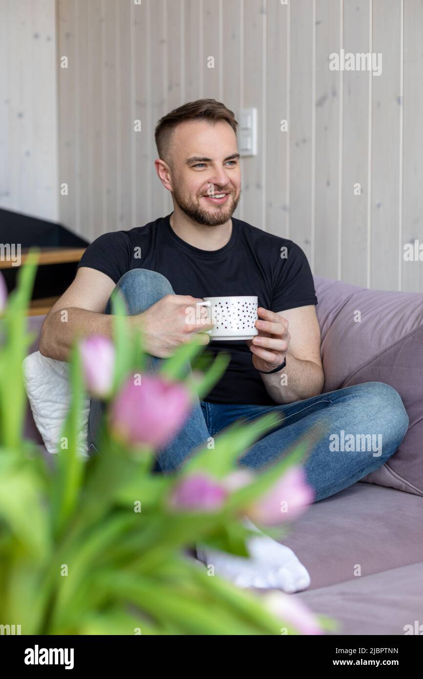 Happy smiling young man in black T-shirt and jeans is sitting on cozy sofa and enjoying cup of hot tea. Rest and relaxation. Happy moments. Positive Stock Photo