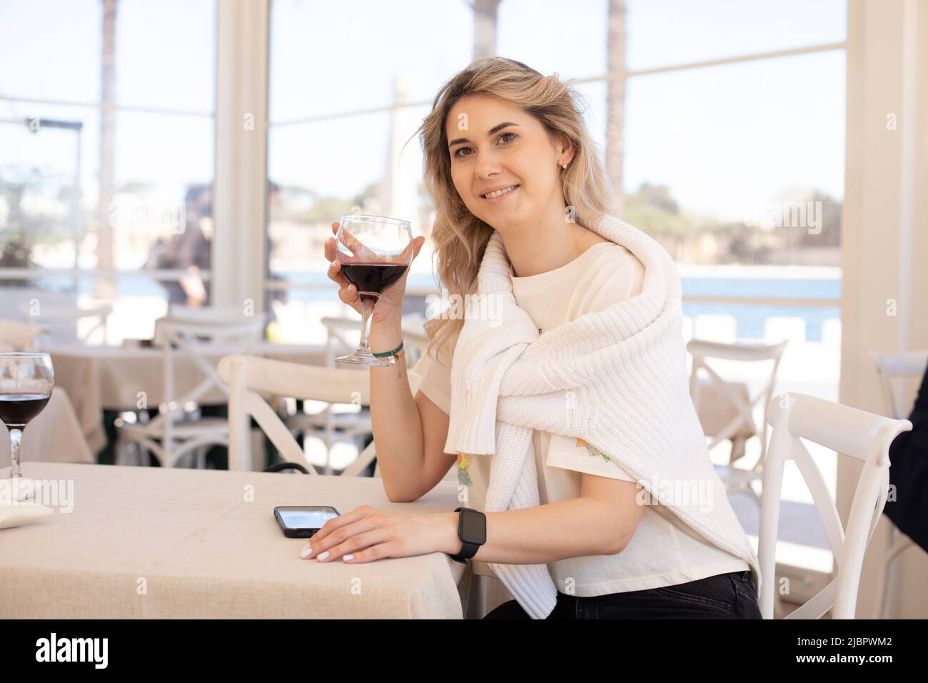 Portrait of young happy woman sitting at table near smartphone, holding glass of wine in restaurant near embankment. Stock Photo