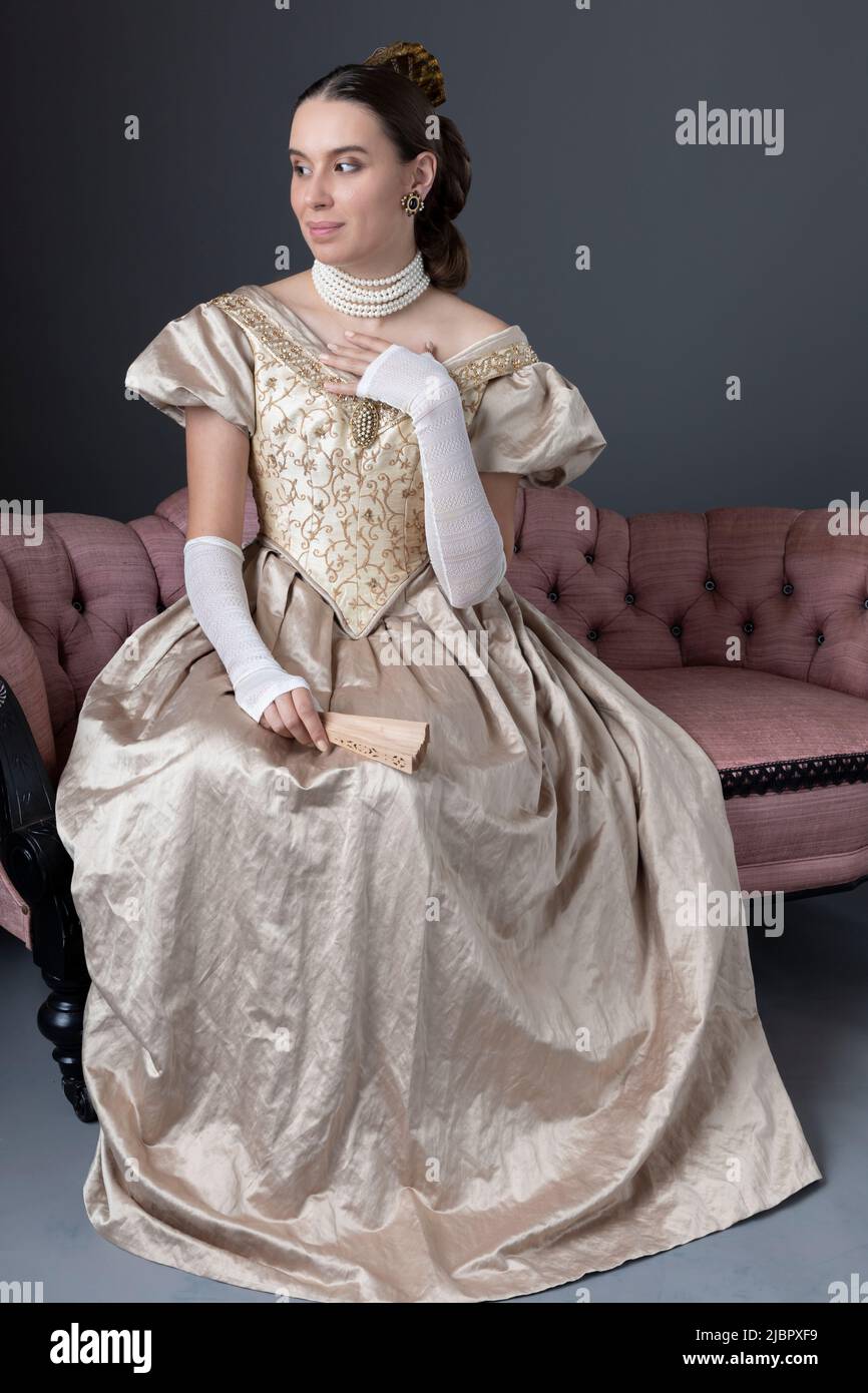 A Victorian woman wearing a gold ball gown and sitting on a pink sofa Stock Photo