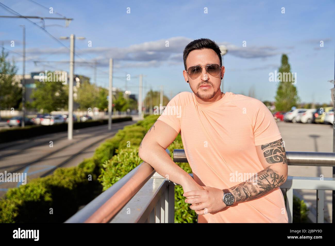 Serious young man, wearing a T-shirt and sunglasses, with a tattoo on his arm, standing on a bridge and looking at the camera on a sunny day on a city street. Stock Photo