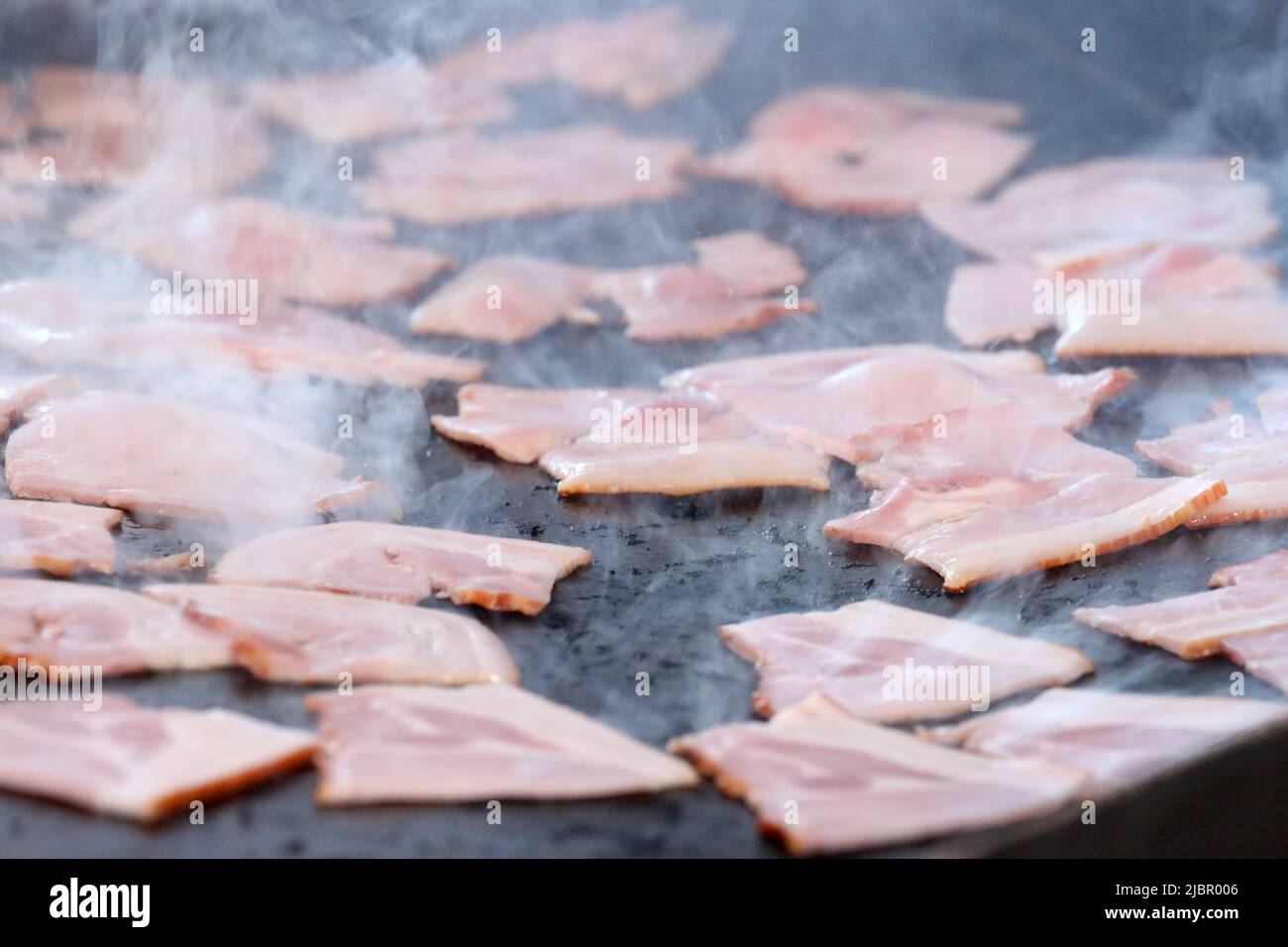 A close up images of a large amount of bacon cooking and sizzling on a black bbq rill or hot plate. Smoke partially blurring the background as the bac Stock Photo