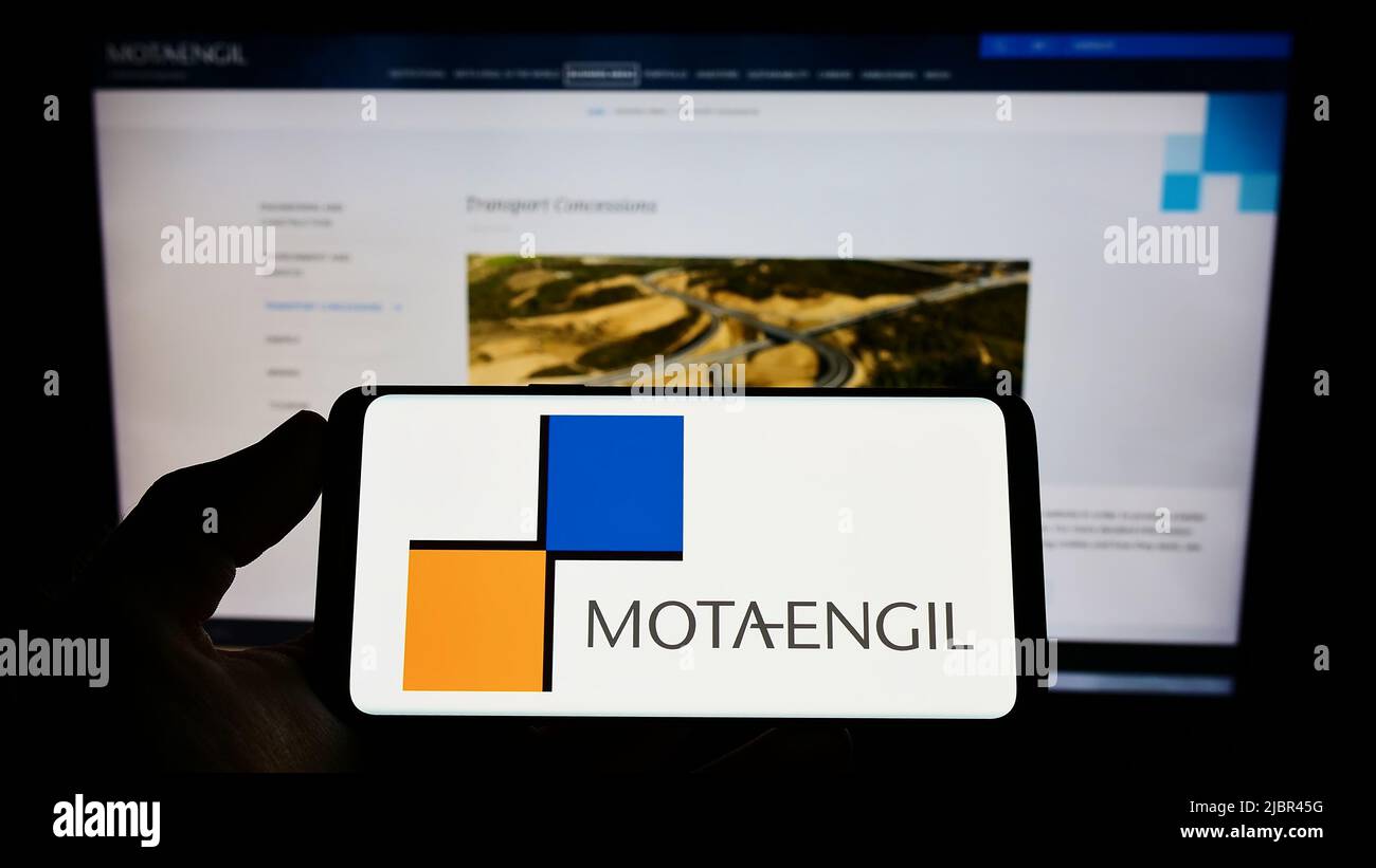 Person holding mobile phone with logo of infrastructure company Mota-Engil SGPS S.A. on screen in front of web page. Focus on phone display. Stock Photo