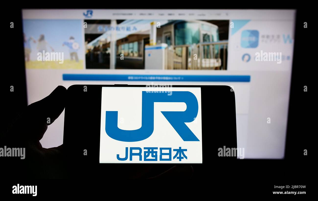 Person holding smartphone with logo of Japanese company West Japan Railway (JR) on screen in front of website. Focus on phone display. Stock Photo