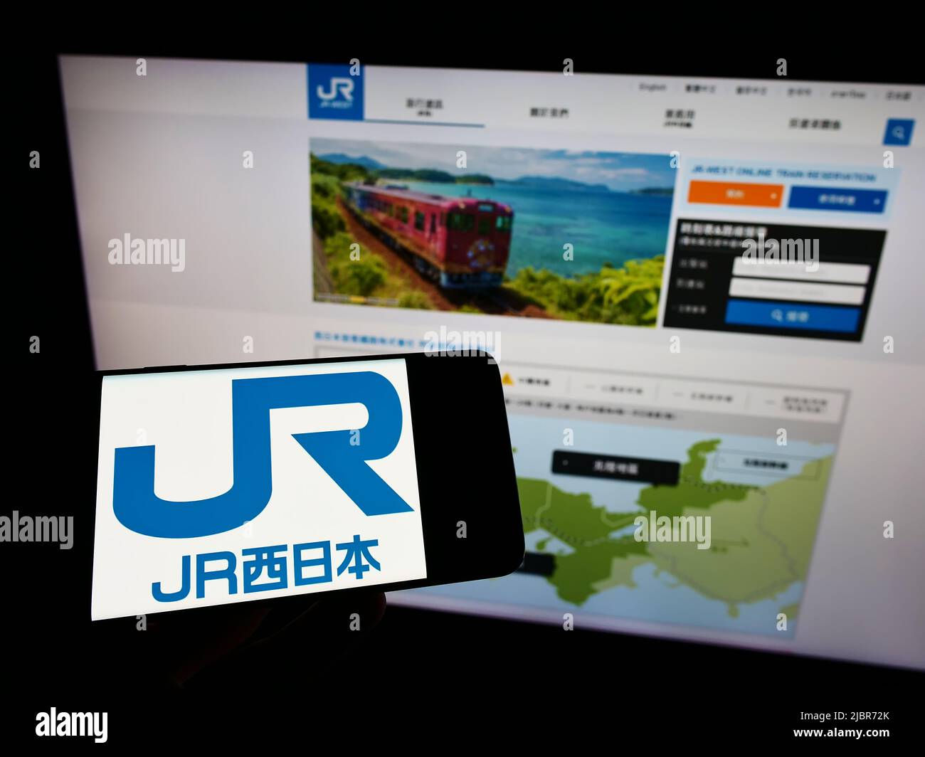 Person holding mobile phone with logo of Japanese company West Japan Railway (JR) on screen in front of business web page. Focus on phone display. Stock Photo
