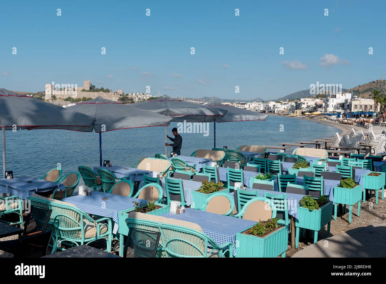 Bodrum, Mugla, Turkey. April 21st 2022 A waiter prepares tables on the beach side cafe in Bodrum harbour with Bodrum Castle in the background, the Aeg Stock Photo