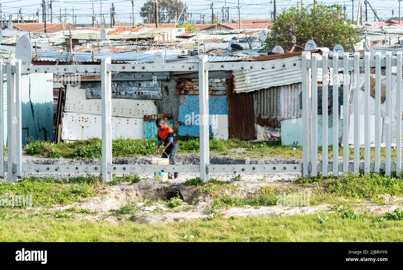 young black African boy carries a bucket in a township against a backdrop of tin shacks or housing concept daily life in South Africa and poverty Stock Photo