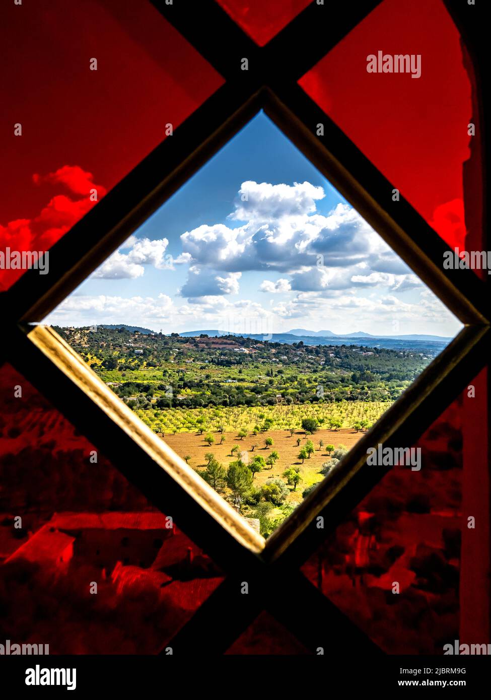 Portrait taken from the Sa Muntanyeta southern slopes of a lush landscape seen through the missing glass in a red colored glass window from the inside Stock Photo