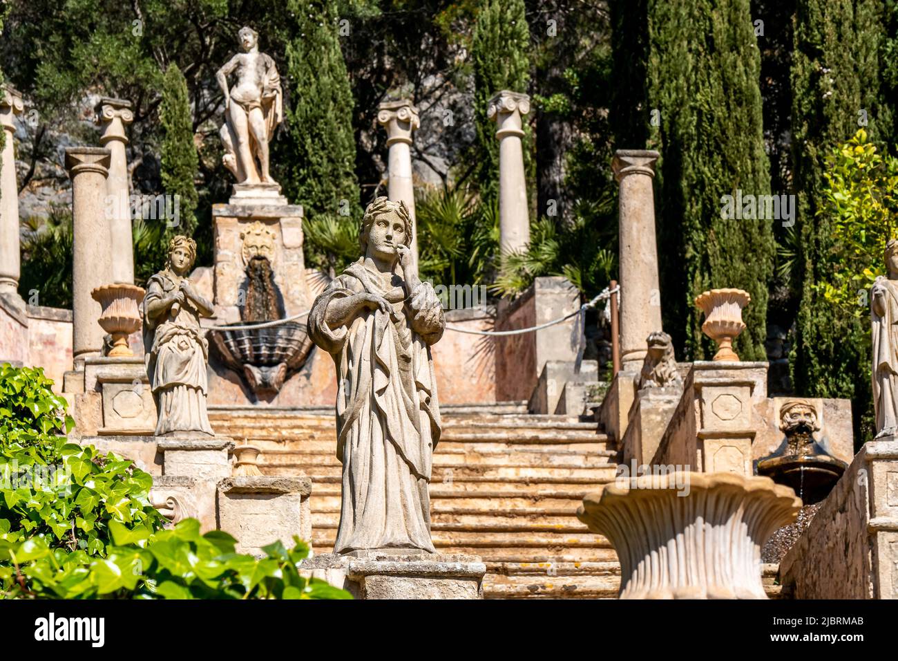 Female statue in front of a staircase called Apollo Staircase lined with gargoyles and amphorae next to cypresses and ivy leads to an Apollo monument. Stock Photo