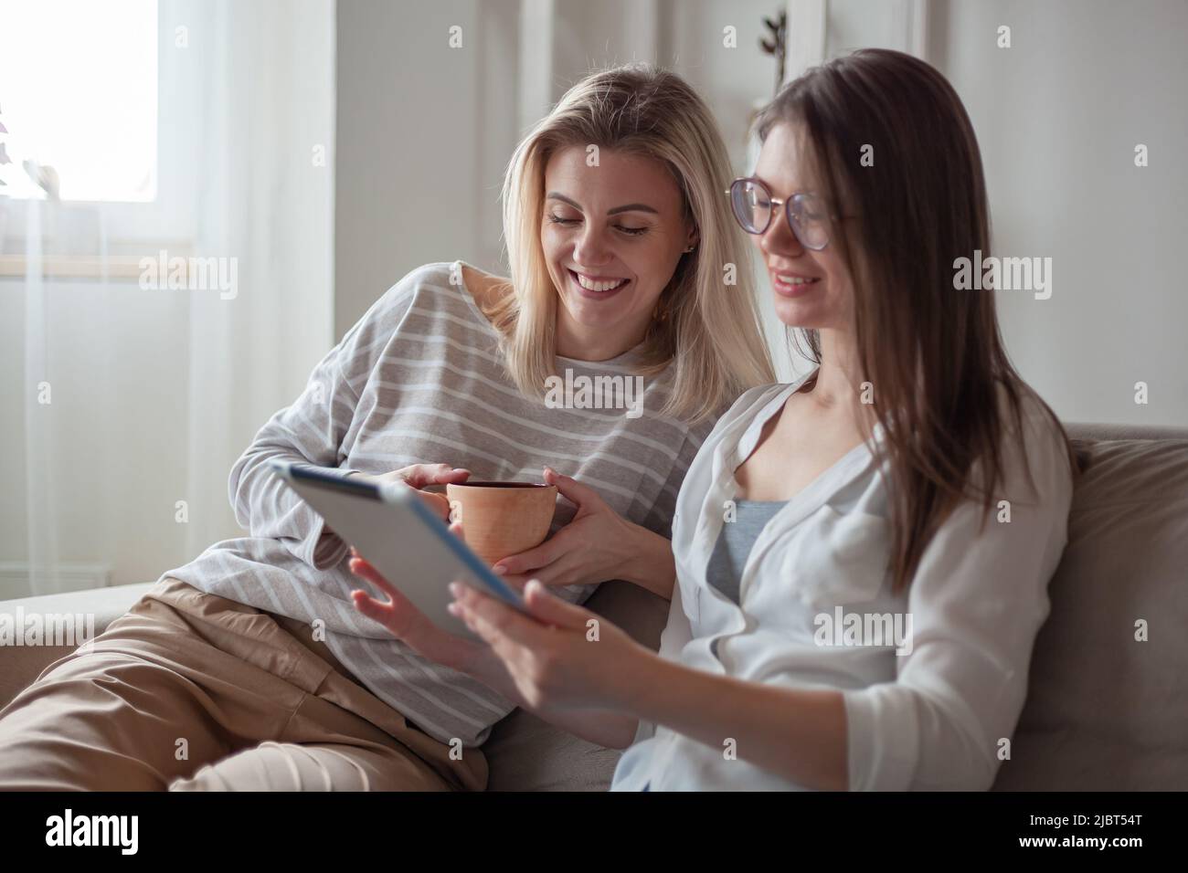 Two young women friends using tablet together, sitting on the couch smiling and fun Stock Photo