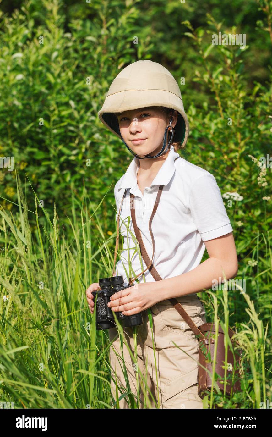 Preteen girl wearing pith helmet stands in the grass holding binoculars observing summer nature. Discovery and adventures concept Stock Photo