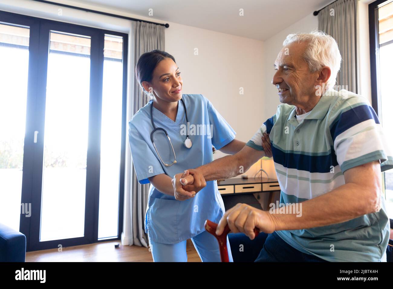 Biracial female physiotherapist assisting caucasian senior man in standing with walking cane at home Stock Photo