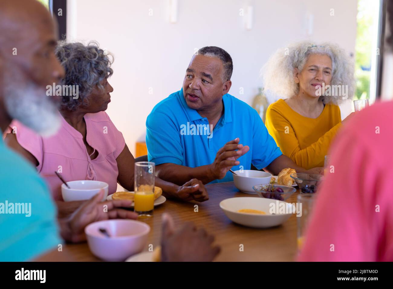 Multiracial senior friends talking and eating healthy breakfast at dining table in nursing home Stock Photo