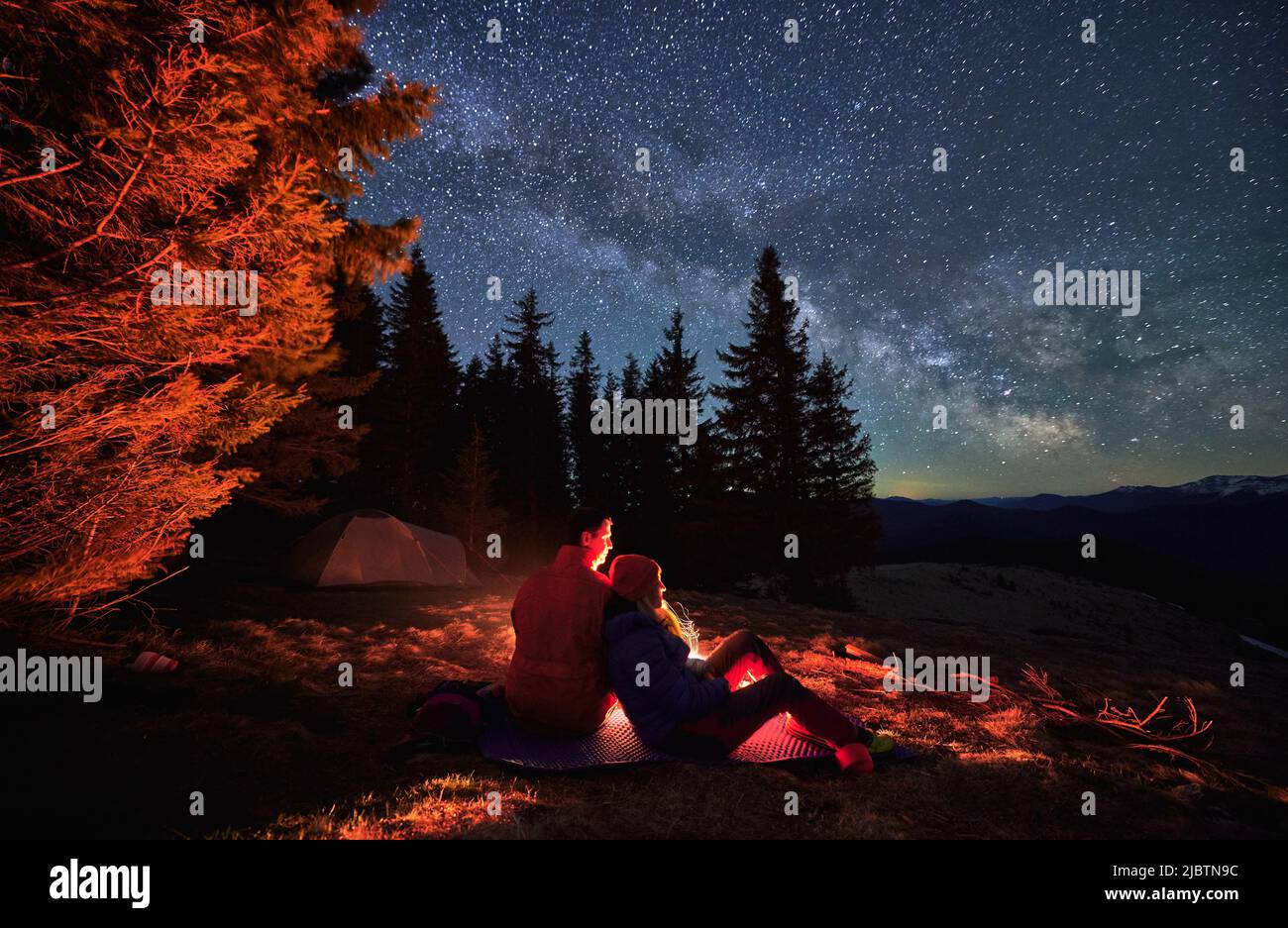 Couple in love sitting on mat, admiring fairytail starry sky with Milky way. Romantic cozy atmosphere in the fresh air against backdrop of tent, trees, mountain hills and millions of stars. Stock Photo