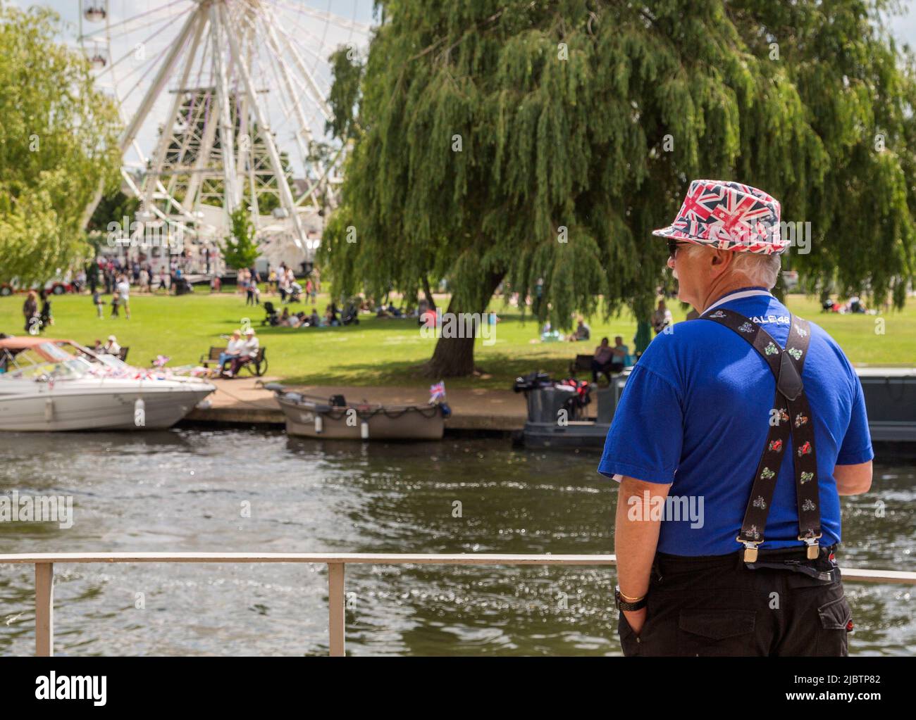 A man in a Union Flag hat looks across a river to people relaxing and  enjoying the sun in a park. British holiday, tourism or travel concept. Stock Photo