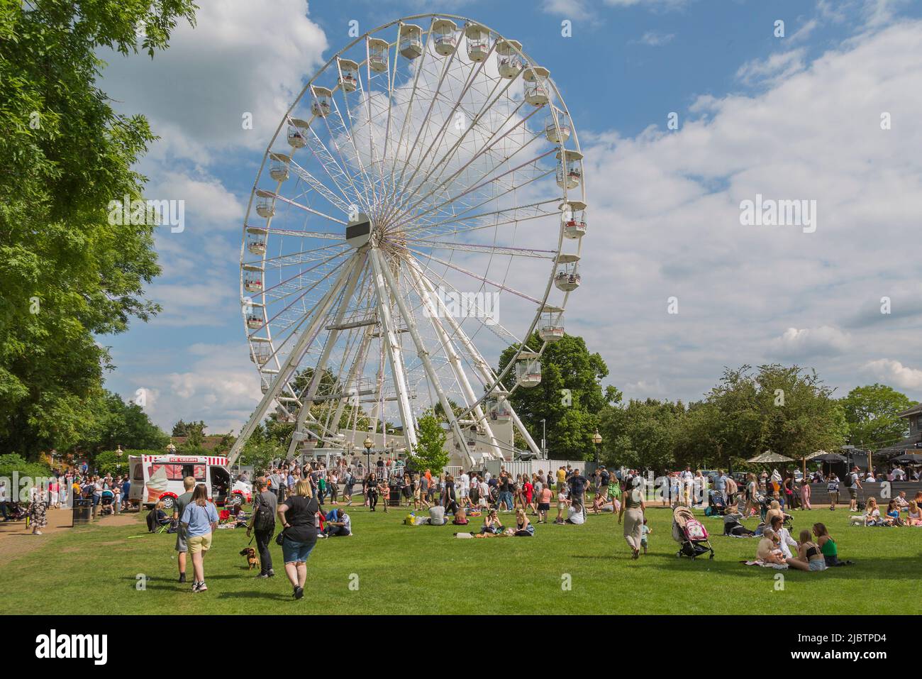 Families enjoy the weather in a park on a sunny summer day, Above them people ride in the big wheel. Stock Photo