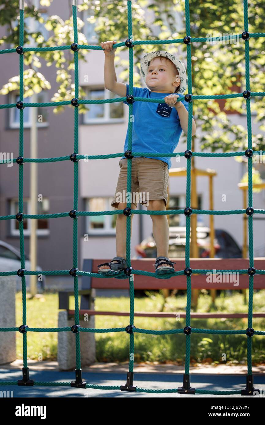 A little boy like a spider climbs up a green net of ropes on a playground. Stock Photo
